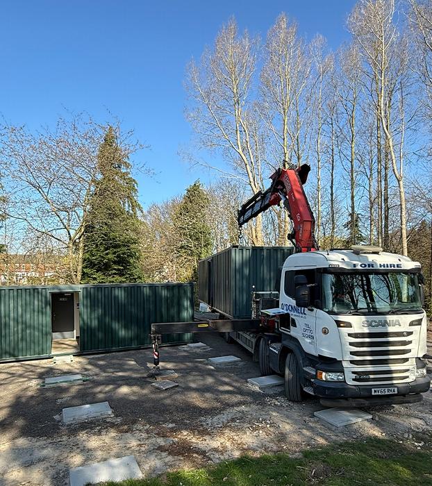 Truck with crane lifting green containers in a construction site surrounded by trees and blue sky showcasing equipment for hire and outdoor work 2 containers being positioned